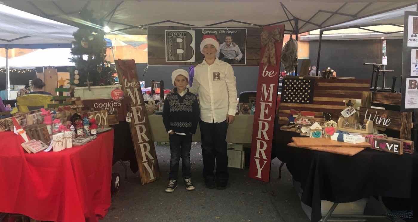 The brothers in Santa hats at a Christmas market booth with handmade wood signs