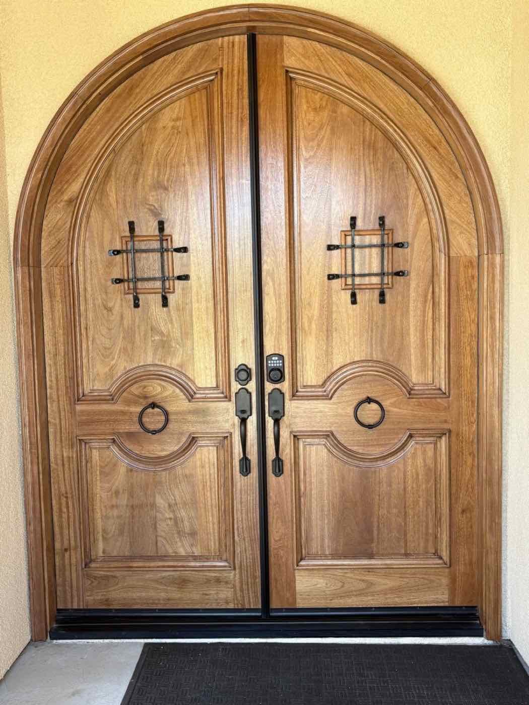 The same arched wood double doors after refinishing, warm natural wood with black iron hardware
